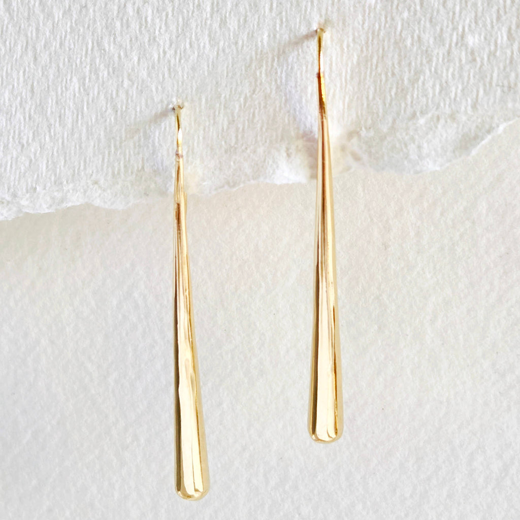 A pair of long, tapered gold dangle earrings with a sleek, smooth finish, hanging against a textured white background.