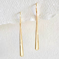 A pair of long, tapered gold dangle earrings with a sleek, smooth finish, hanging against a textured white background.