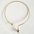 A minimalist brass collar necklace with a polished brass ball at each end, photographed on a textured off-white background.