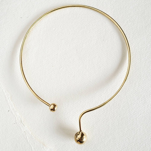 A minimalist brass collar necklace with a polished brass ball at each end, photographed on a textured off-white background.