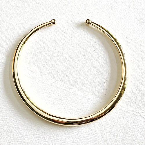 A polished gold hoop collar necklace with a small ball detail at each end, displayed on a textured white fabric background.