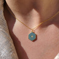 A close-up of a person's neck and chest wearing a delicate gold chain necklace with a hexagonal pendant. The pendant features a small opal star design at the center.
