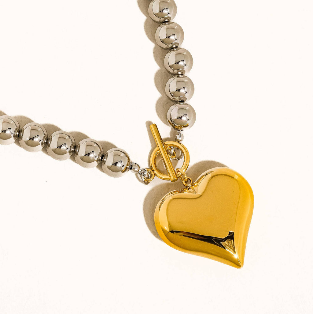 A close-up of a necklace featuring a polished gold heart pendant and a silver-toned beaded chain with a toggle clasp, set against a clean off-white background.