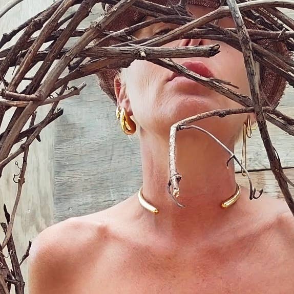A close-up lifestyle shot of a woman wearing gold jewelry, including a gold collar necklace, gold earrings, with dried branches framing her face against a rustic wooden background.