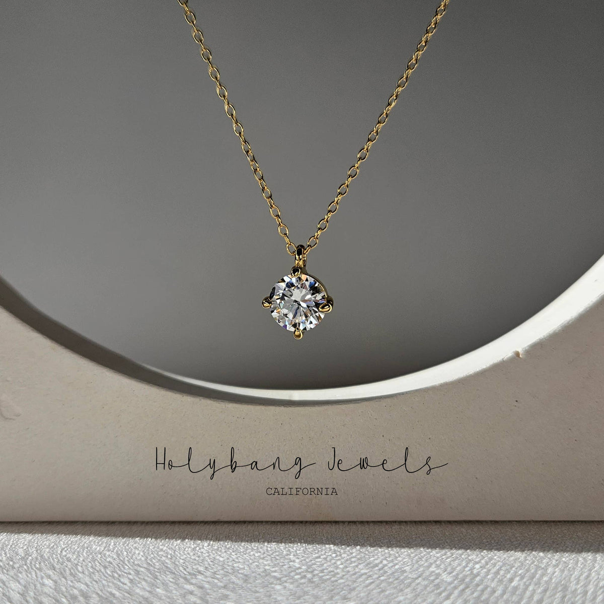 A gold necklace with a clear gemstone pendant displayed on a beige jewelry box. The box has the brand name 'Holybang Jewels' printed on it.