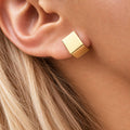 A close-up of a woman's ear wearing a small geometric gold stud earring with a faceted cube design.
